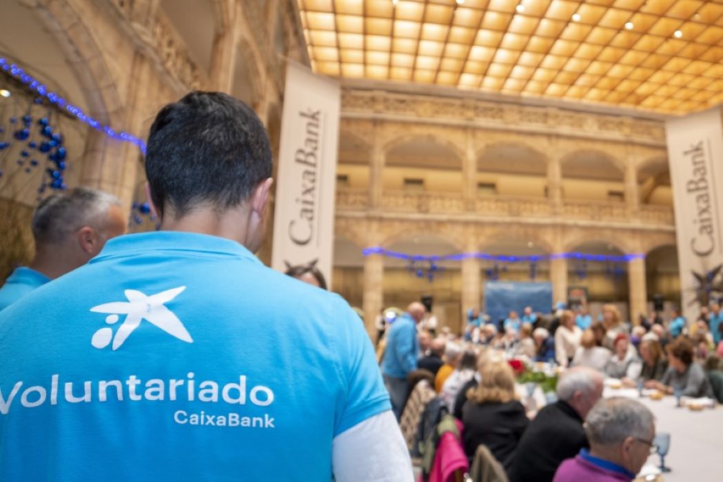 Persona amb samarreta blava del programa de voluntariat de CaixaBank en un esdeveniment multitudinari en un edifici hist&ograve;ric amb pancartes de l&rsquo;entitat.