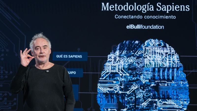 Ferran Adri&agrave; en un moment de la presentaci&oacute;, davant d&rsquo;una pantalla blava on es llegeix &lsquo;Metodologia Sapiens&rsquo; sobre la representaci&oacute; d&rsquo;un cervell.