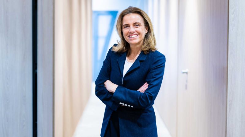 Ana Mart&iacute;n de Santa Olalla, amb un vestit blau mar&iacute; i els bra&ccedil;os creuats, posant en un passad&iacute;s d&rsquo;oficines amb parets de fusta clara.