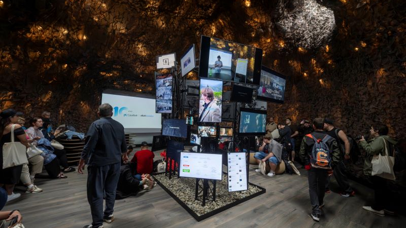 Experiència immersiva entre pantalles Interior d'una sala de museu com una cova. Al centre hi ha una estructura vertical formada per moltes pantalles que mostren vídeos, imatges i textos diferents.