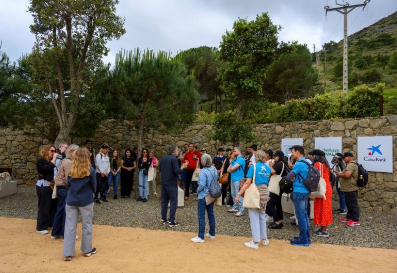 Grup de persones a l'exterior d'elBulli1846. Grup de persones reunides a l'aire lliure, escoltant en Lluís García que parla al centre. Estan en un entorn natural amb arbres i es veu un mur de pedra i cartells d'empreses col·laboradores.