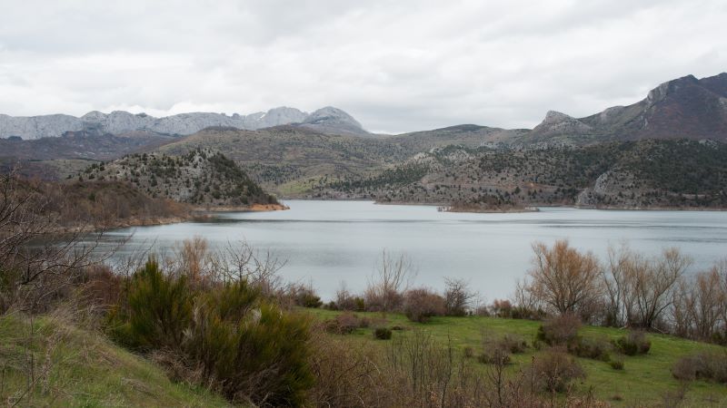 Caldas de Luna: aigües termals en plena Reserva de la Biosfera Embassament envoltat de muntanyes i turons amb vegetació dispersa sota cel ennuvolat, amb ribes verdes i arbres sense fulles en primer pla