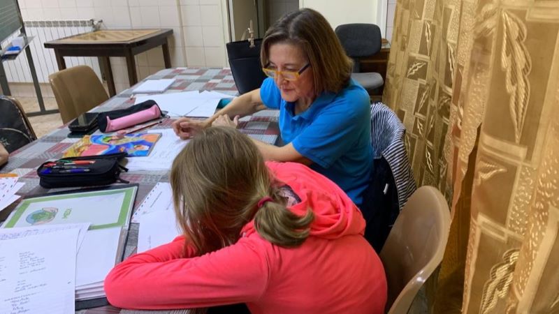 Teresa Vázquez, prejubilada com a directora d’oficina bancària, acompanya infants en un programa de reforç escolar. Persona voluntària acompanya una estudiant en una activitat educativa, revisant exercicis i materials d’aprenentatge en una taula.