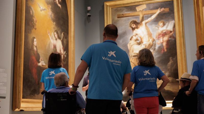 Voluntariat CaixaBank i Apropa acosten a la gent gran la vida cultural de les seves ciutats Voluntariat acompanya persones usuàries de cadira de rodes durant una visita guiada en un museu, observant grans pintures exposades a la sala.