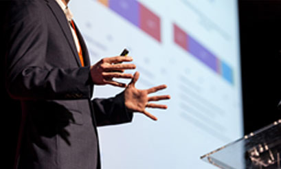 Persona vestida con traje y corbata, haciendo una presentación. En el fondo se ve una proyección de diapositivas con gráficas y texto.