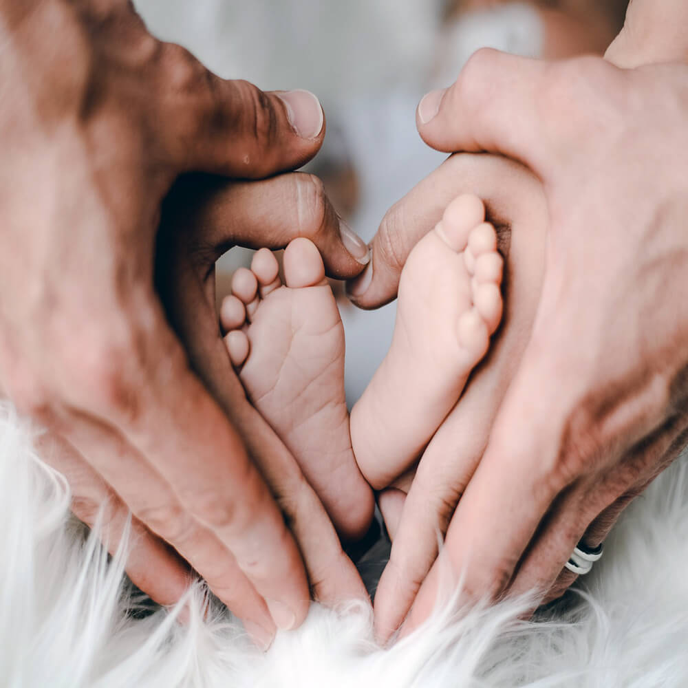 Manos de unos padres formando un corazón mientas sujetan los pies de su hijo