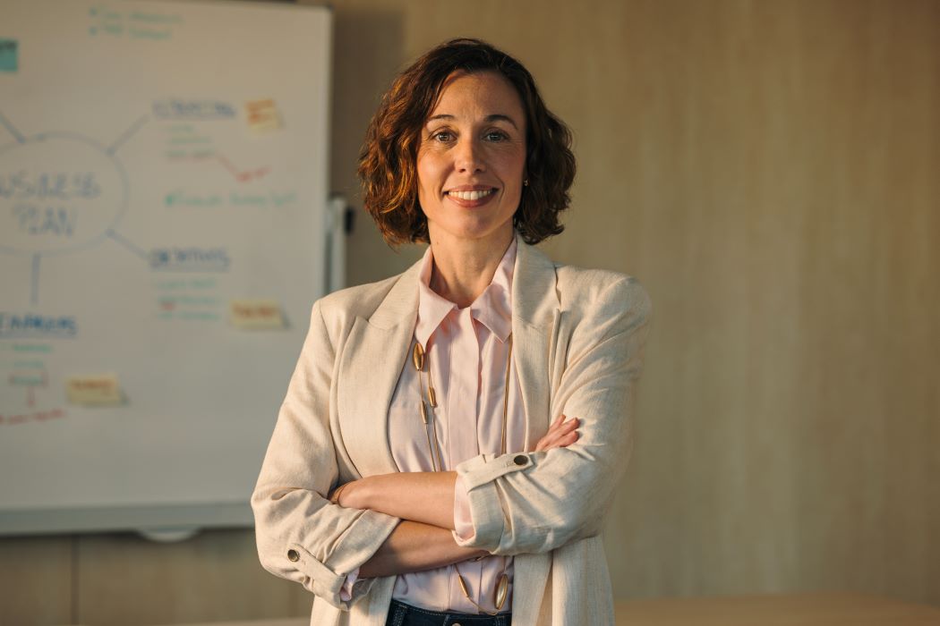 Dona amb els braços plegats davant d'una pissarra amb esquemes en un entorn professional.