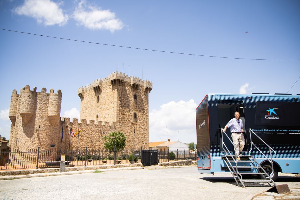 Un hombre desciende de un ofimóvil de CaixaBank estacionado junto a un castillo medieval en un día soleado