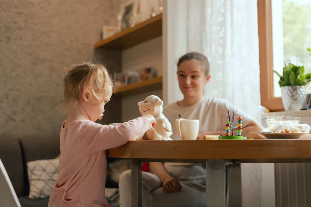 Una nena de menys de tres anys juga amb un peluix mentre esmorza amb la seva mare
