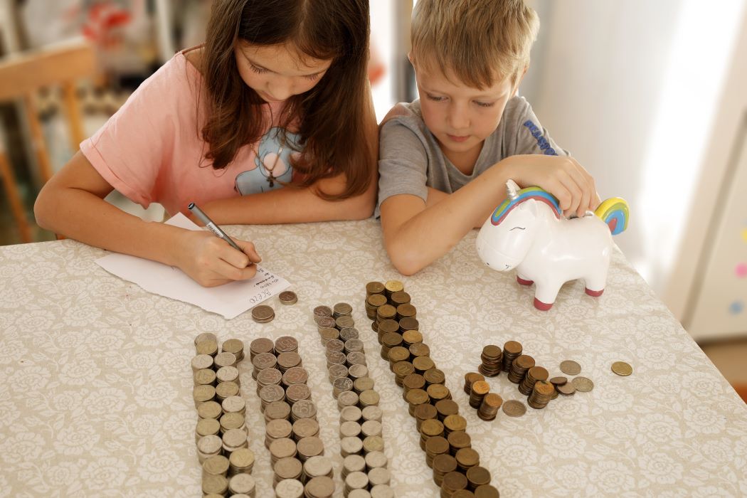 Dos niños de entre seis y diez años cuentan monedas que tienen acumuladas en montones y hacen cuentas sobre sus ahorros