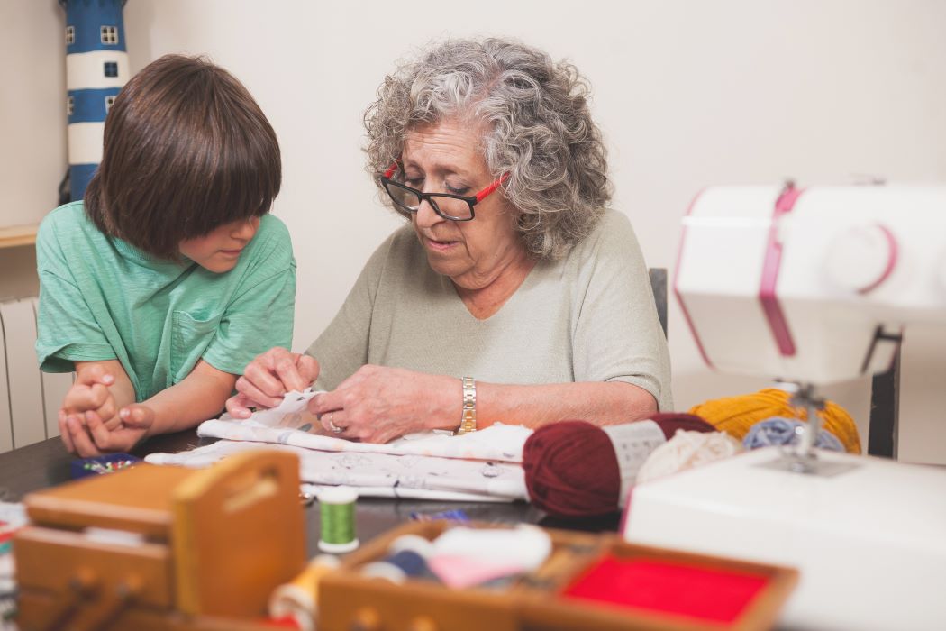 Persona mayor enseñando a un niño a coser sobre una mesa con materiales de costura y una máquina de coser