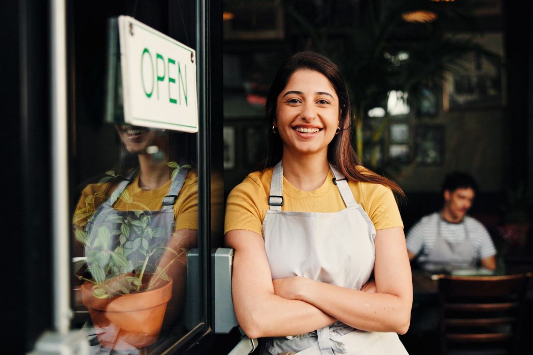 Trabajadora con delantal de pie en la entrada de un local con cartel de abierto