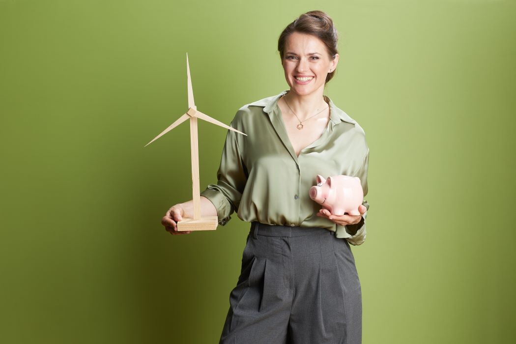 Mujer de pie sosteniendo un modelo de aerogenerador en una mano y una hucha rosa en la otra, sobre un fondo verde.