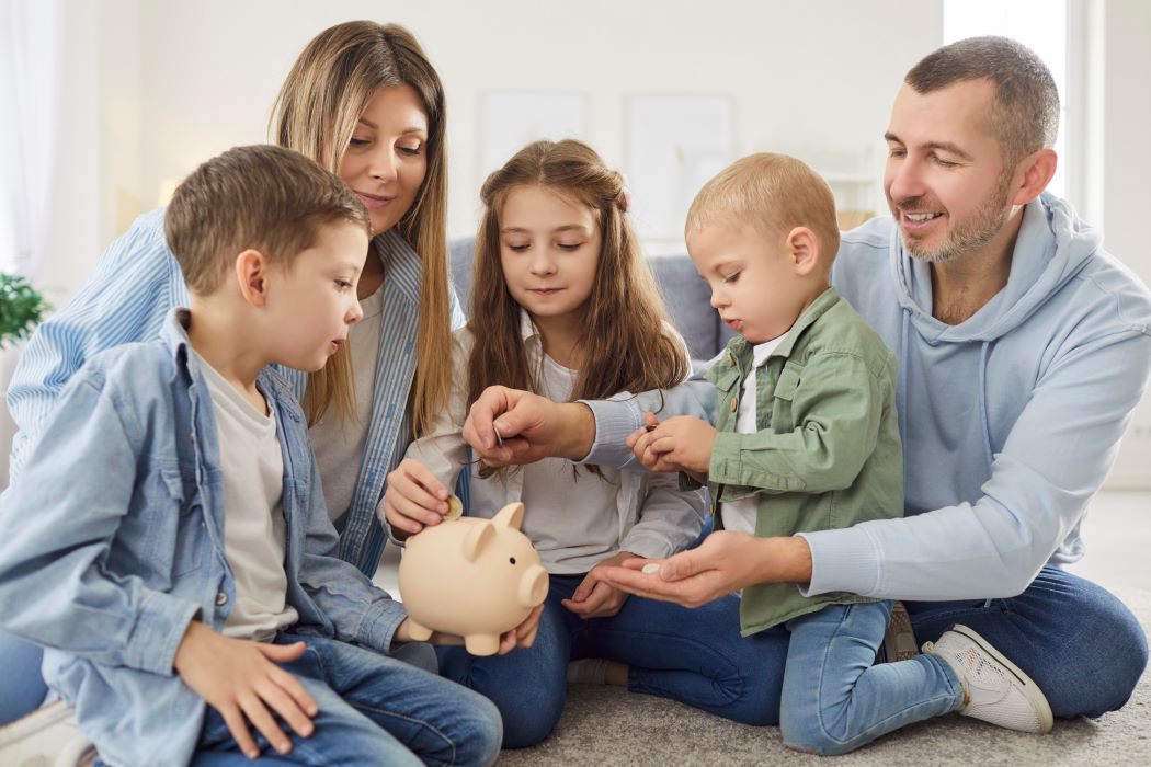 Una família asseguda a terra introdueix monedes en una guardiola amb forma de porquet mentre comparteixen un moment d'estalvi conjunt.