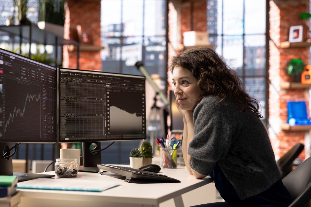 Mujer trabajando en una oficina moderna frente a dos monitores con gráficos y datos financieros