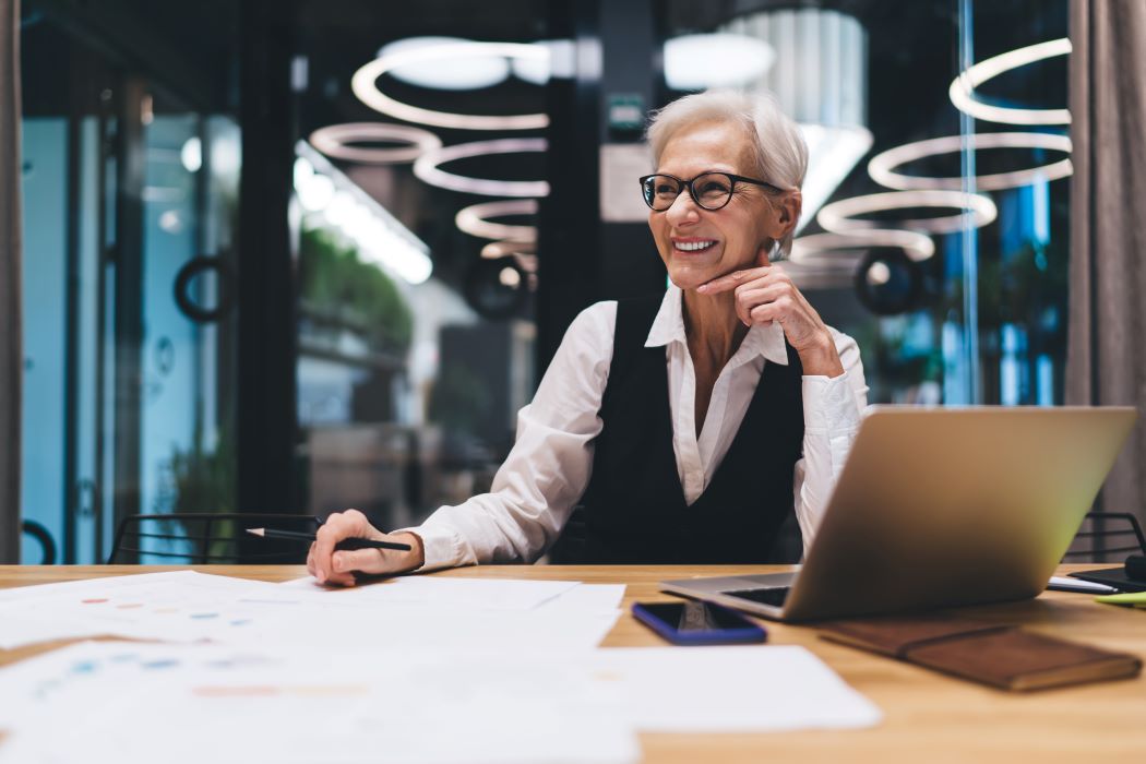 Dona sènior revisant documents sobre una taula en un entorn d’oficina modern, amb un ordinador portàtil obert i material de treball distribuït al davant