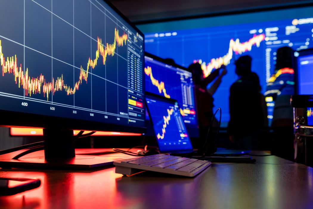 Pantallas de trading con gráficos financieros en alza y varias personas analizando los datos proyectados al fondo.