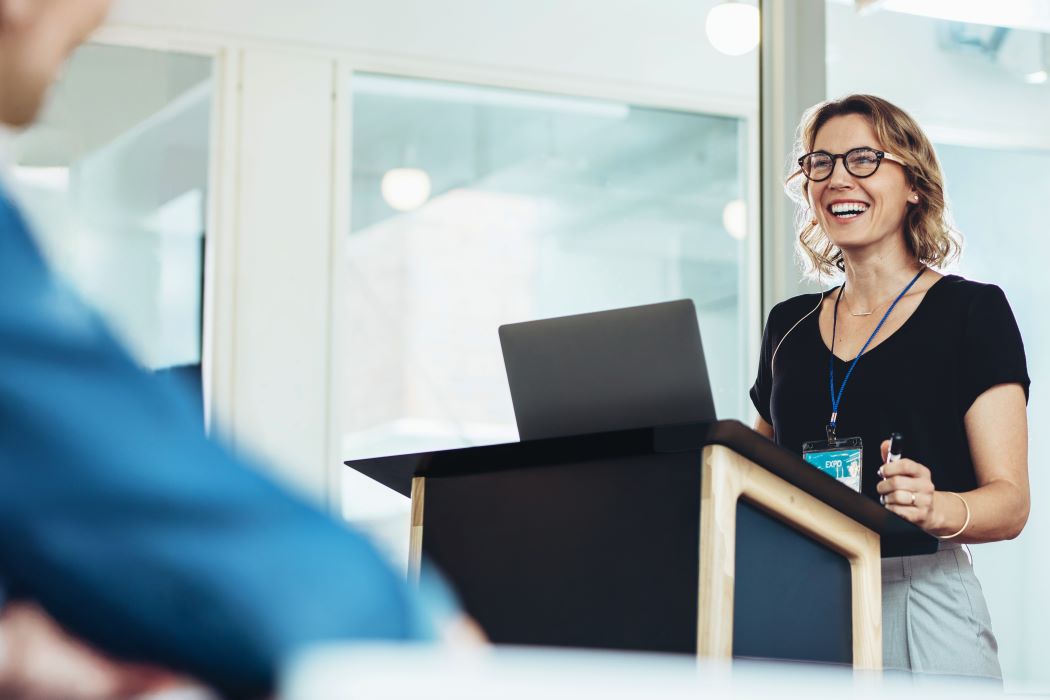 Dona directiva parlant des d’un faristol en un espai professional modern, amb una presentació davant d’una audiència en un entorn corporatiu