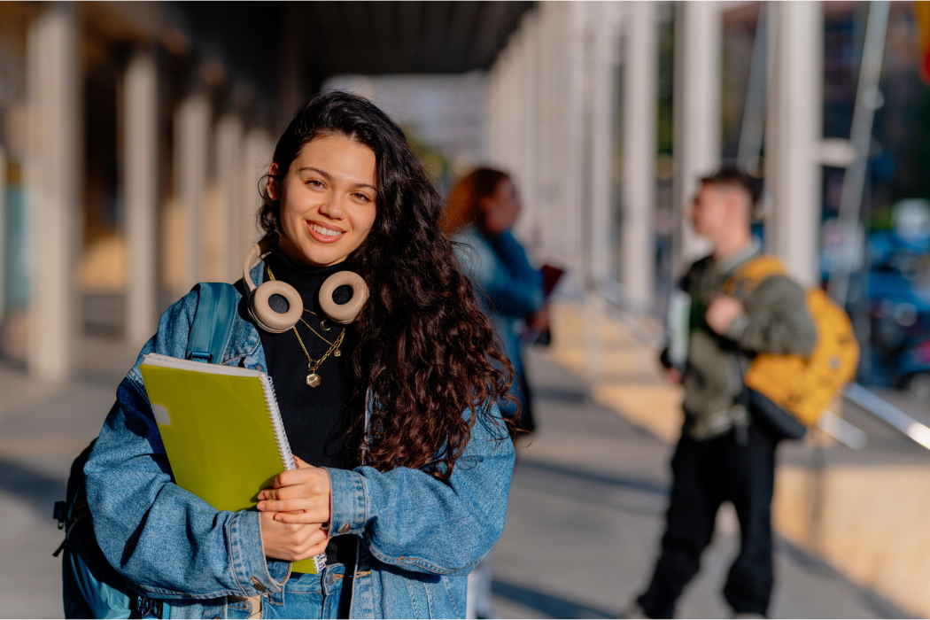 Estudiante universitaria mira a cámara con un cuaderno amarillo y cascos.