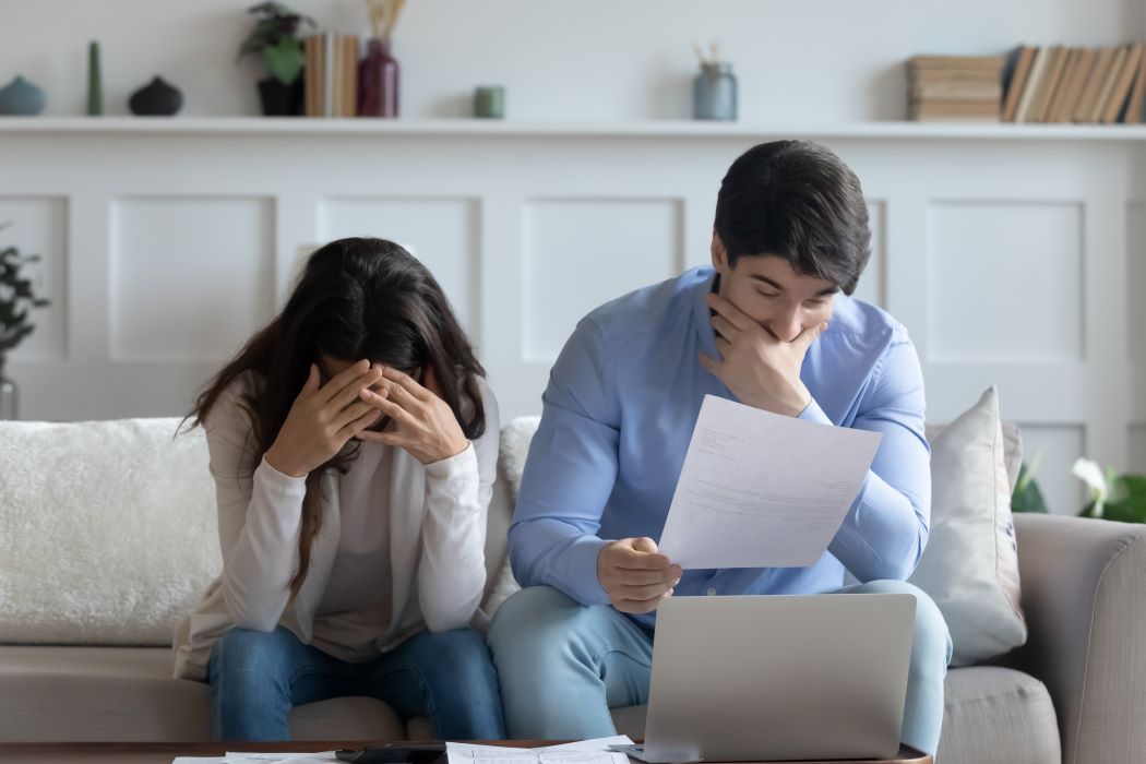 Dues persones preocupades assegudes en un sofà revisen documents a casa al costat d'un ordinador portàtil.
