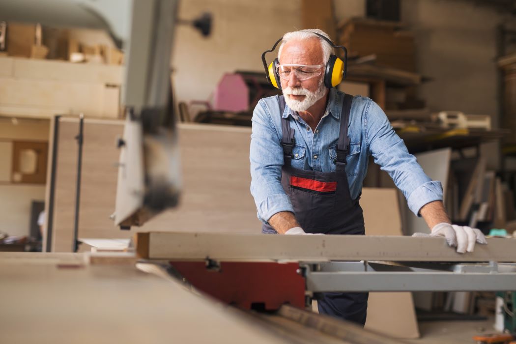 Persona sénior trabaja la madera en un taller, utilizando una sierra de mesa y llevando protección auditiva y guantes