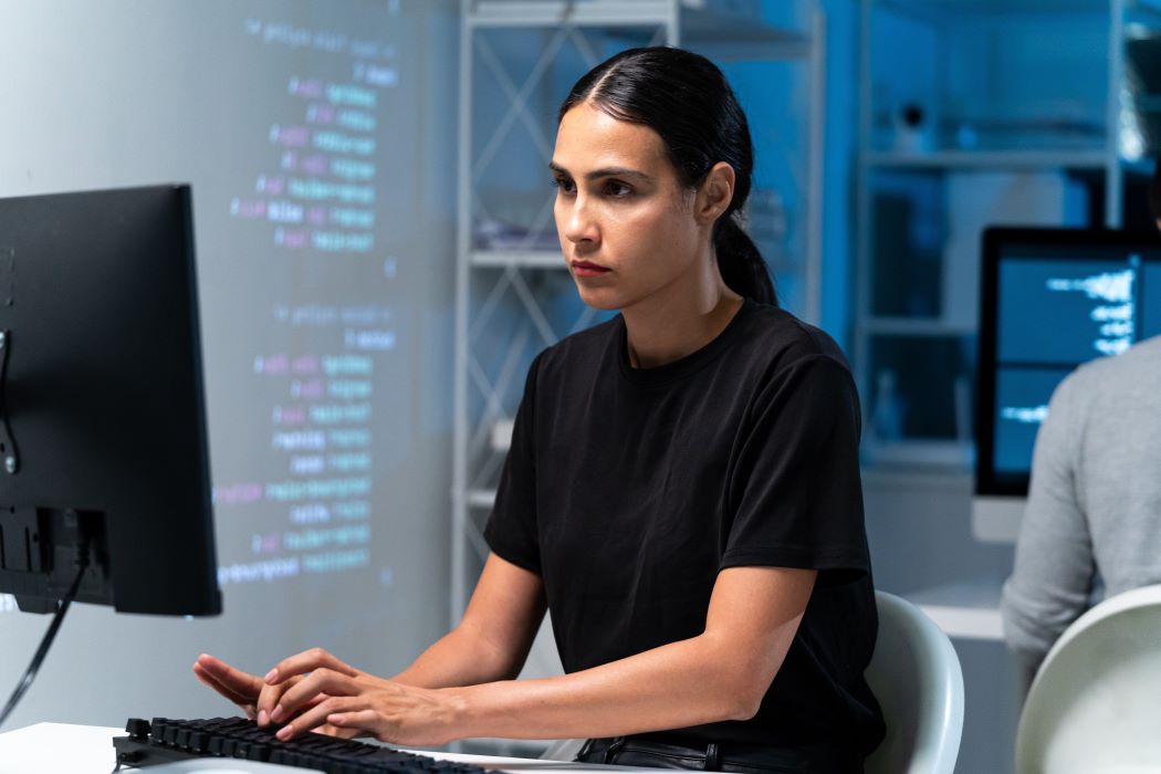 Mujer trabajando con un ordenador en un entorno de formación tecnológica, con pantallas y código informático proyectado al fondo