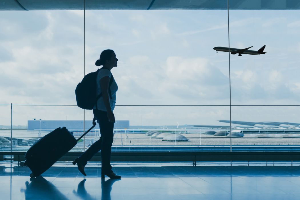 Una mujer pasea por un aeropuerto con una mochila en la espalda y una maleta con ruedas. De fondo se ve un avión despegando