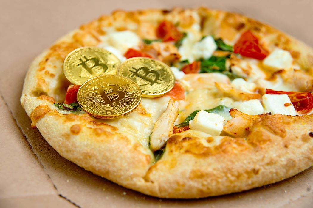 Pizza acabada de fer amb formatge fos, tomàquet i fulles verdes sobre una caixa de cartró; en primer pla, tres monedes daurades amb el símbol de bitcoin col·locades damunt de la pizza