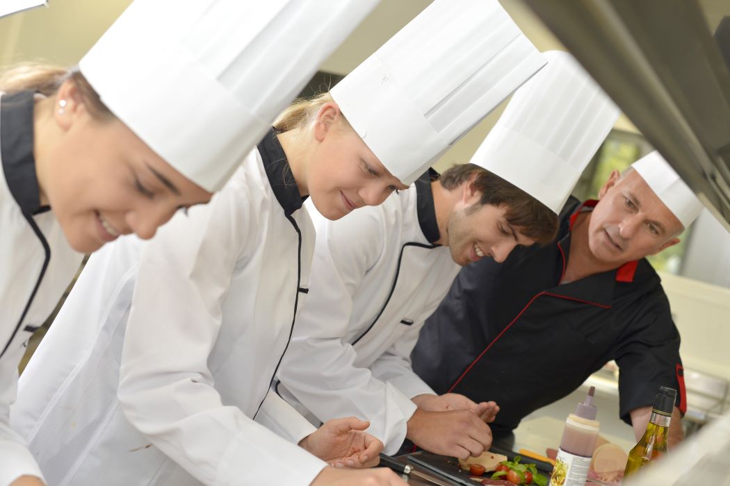 Grup d’estudiants de cuina amb barrets de xef i uniformes, preparant aliments en una cuina professional amb una persona instructora.