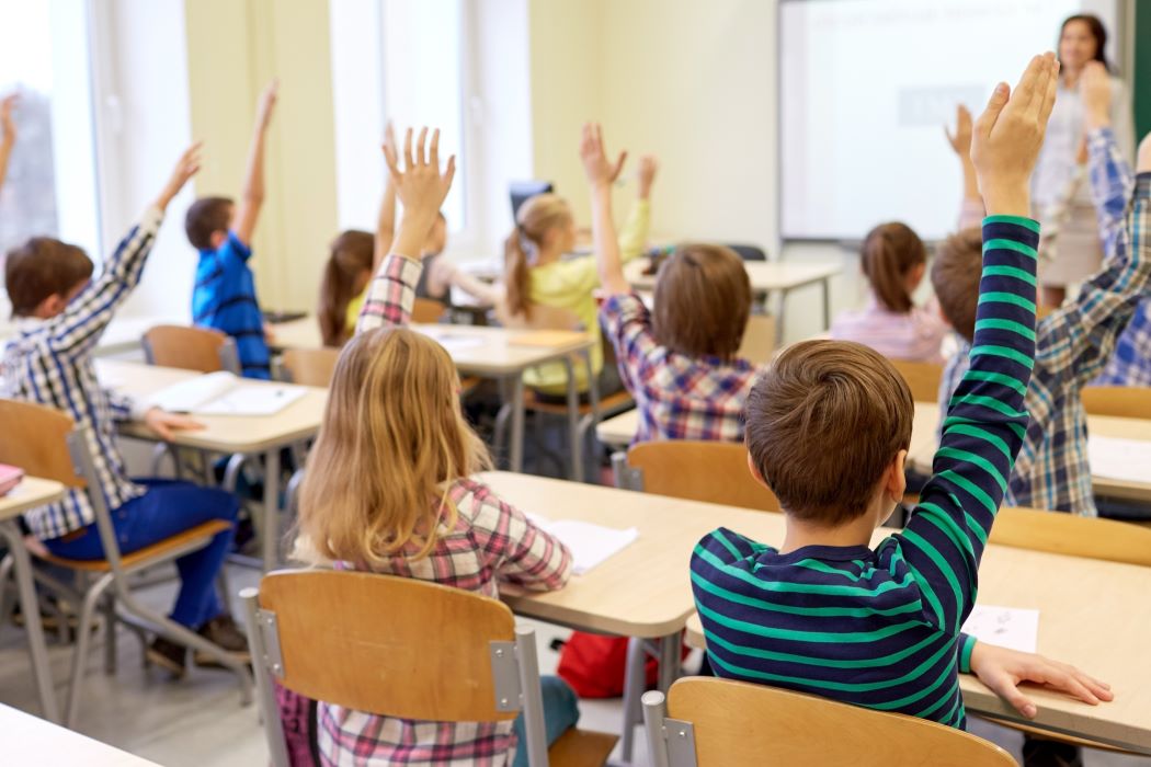 Infants en un aula aixecant la mà mentre participen en una classe amb una persona docent al davant