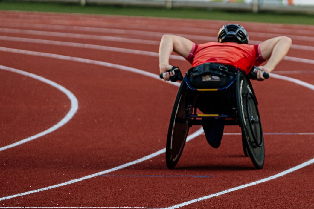 Persona en silla de ruedas deportiva impulsándose con fuerza en una pista de atletismo, durante una carrera en un estadio al aire libre.