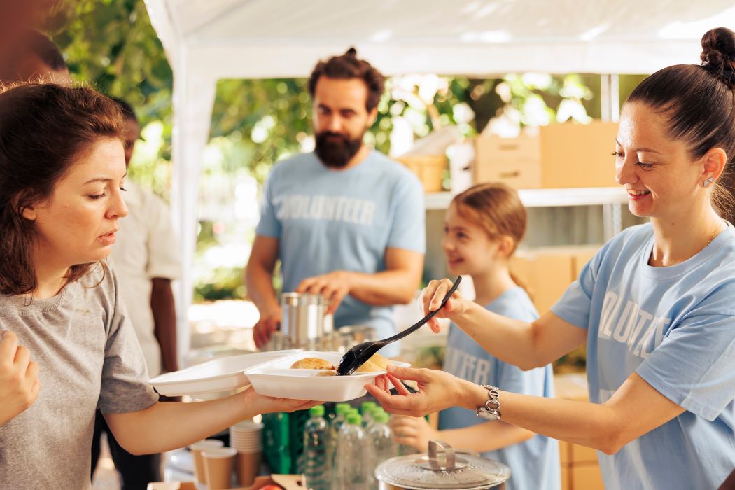 Una familia voluntaria reparte comida caliente en envases biodegradables a una persona en un puesto solidario al aire libre, con alimentos y botellas en una mesa y más voluntarios al fondo.
