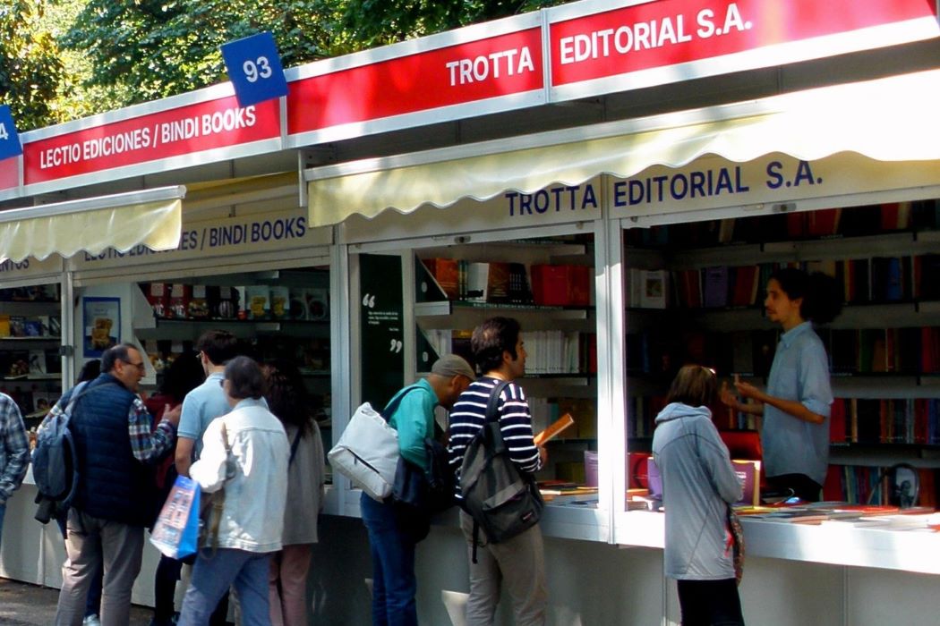 Fila de casetas de la Feria del Libro en un paseo arbolado, con números identificativos y varias personas caminando y consultando libros frente a los puestos