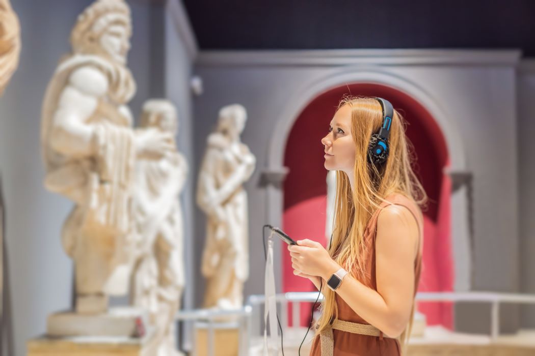 Persona amb auriculars observa escultures clàssiques en un museu mentre sosté una audioguia, amb exposició il·luminada i fons arquitectònic.