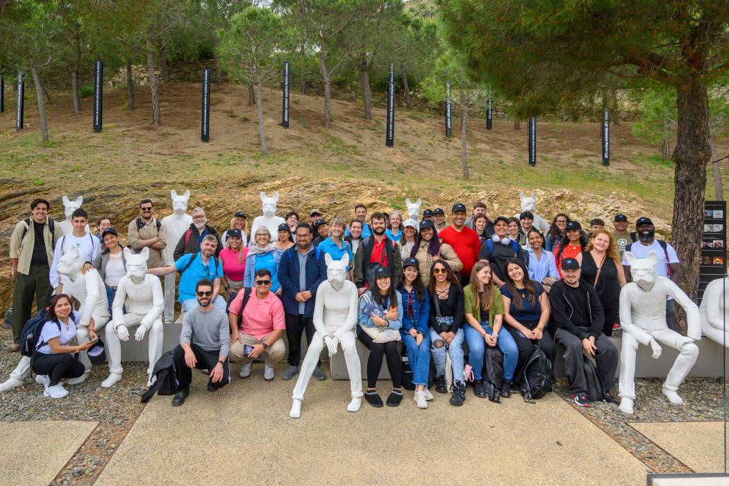 Grupo grande de personas posan para una foto al aire libre. Están sentados entre esculturas blancas de figuras humanas con cabezas de animales, con un fondo de árboles y naturaleza.