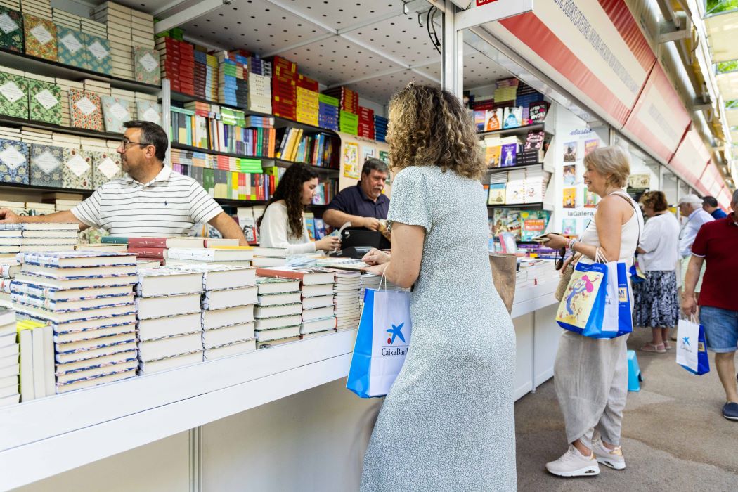 Puesto de una feria del libro al aire libre con varias personas comprando, mientras otras esperan con bolsas reutilizables.