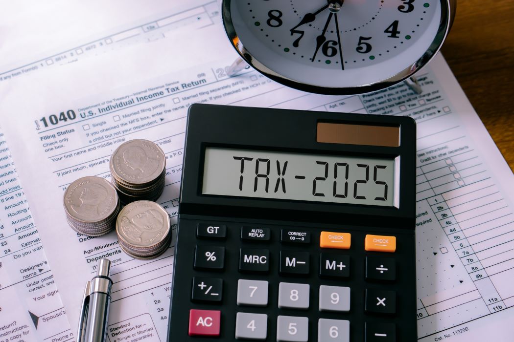 Calculadora con el texto “TAX-2025”, monedas y bolígrafo sobre un formulario fiscal, con un reloj encima que sugiere plazo de declaración de la renta
