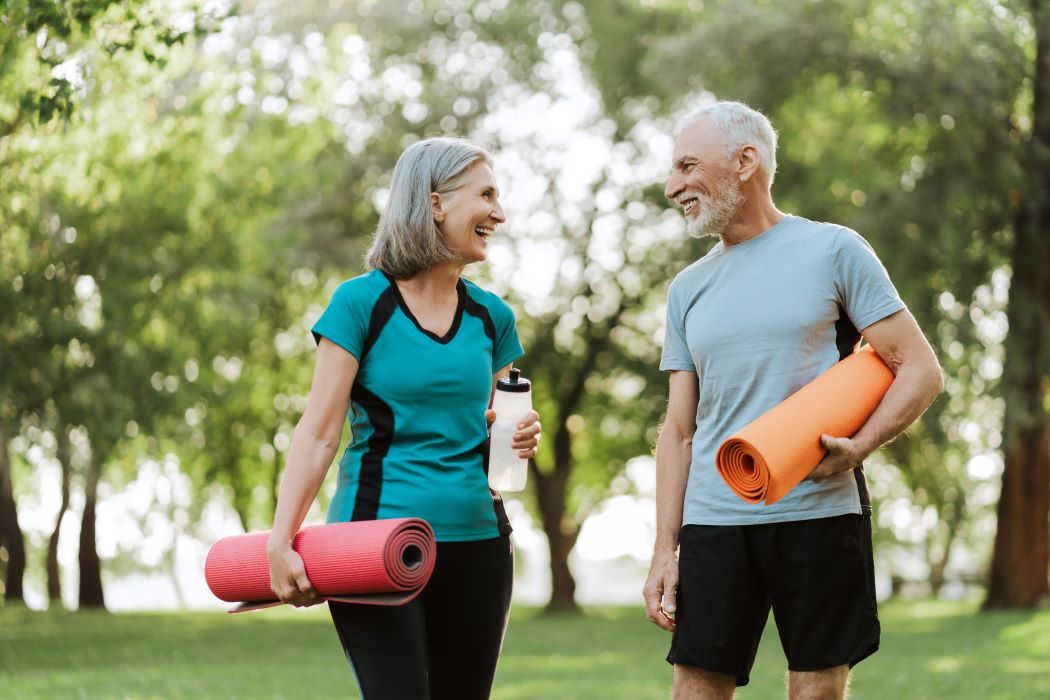 Dos personas adultas mayores conversan en un parque con esterillas de yoga y botella de agua, rodeadas de árboles y luz natural.