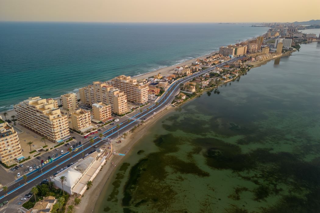 Vista aèria del litoral del Mar Menor amb edificis al costat de la costa, carretera paral·lela i aigües tranquil·les en contrast amb el mar obert