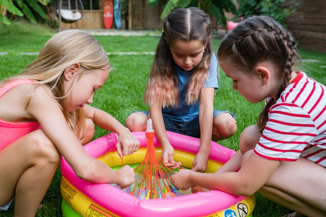 Tres nenes juguen sobre la gespa al voltant d’una petita piscina inflable, omplint globus amb un dispositiu multicolor a l’aire lliure