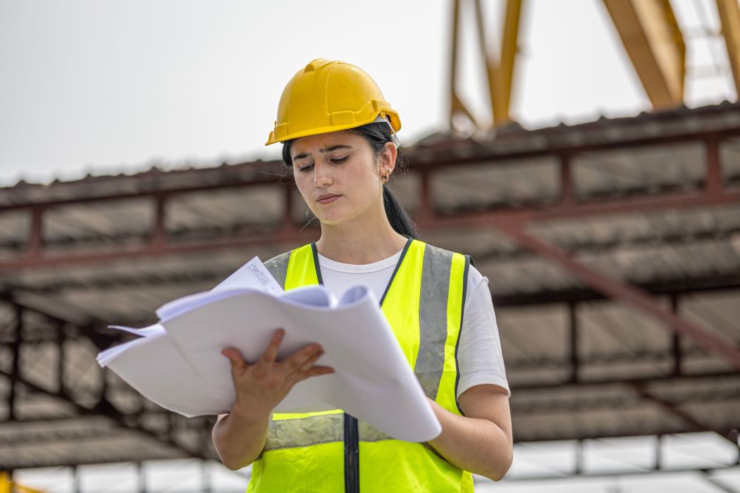 Mujer joven con casco de seguridad y chaleco reflectante revisando planos en una obra de construcción con estructura metálica al fondo.