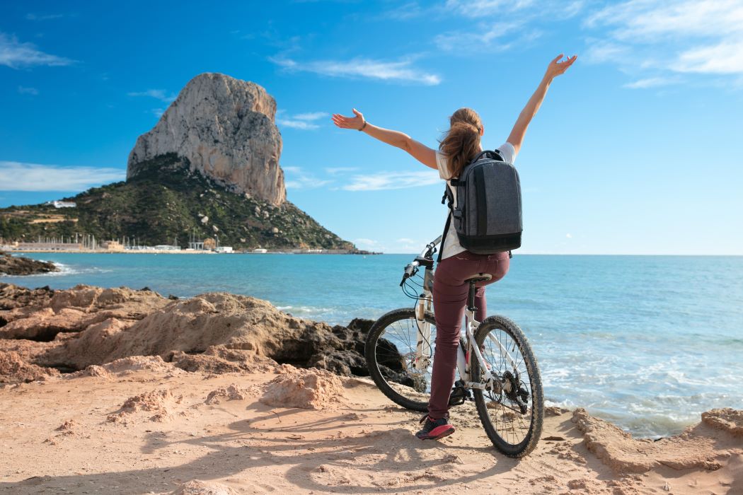 Mujer con bicicleta en la costa rocosa junto al mar, con el Peñón de Ifach al fondo y cielo azul despejado