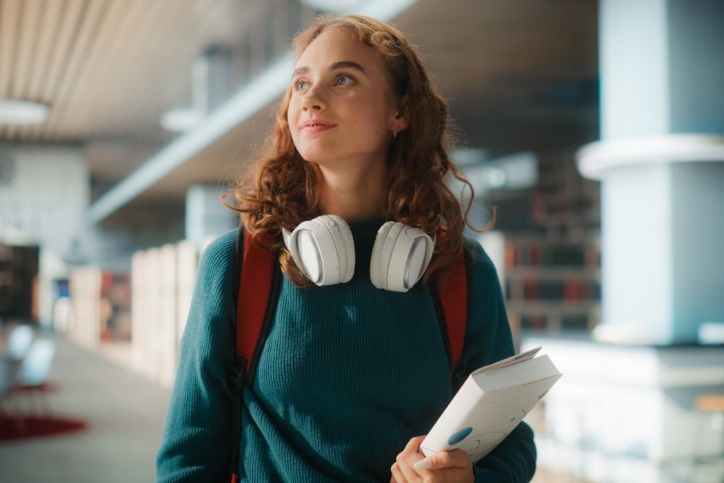 Persona en biblioteca moderna con auriculares al cuello y mochila, sosteniendo un libro mientras recorre el espacio luminoso