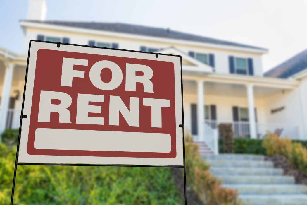 Cartel de “For Rent” en primer plano delante de una casa unifamiliar con porche y jardín desenfocado al fondo.