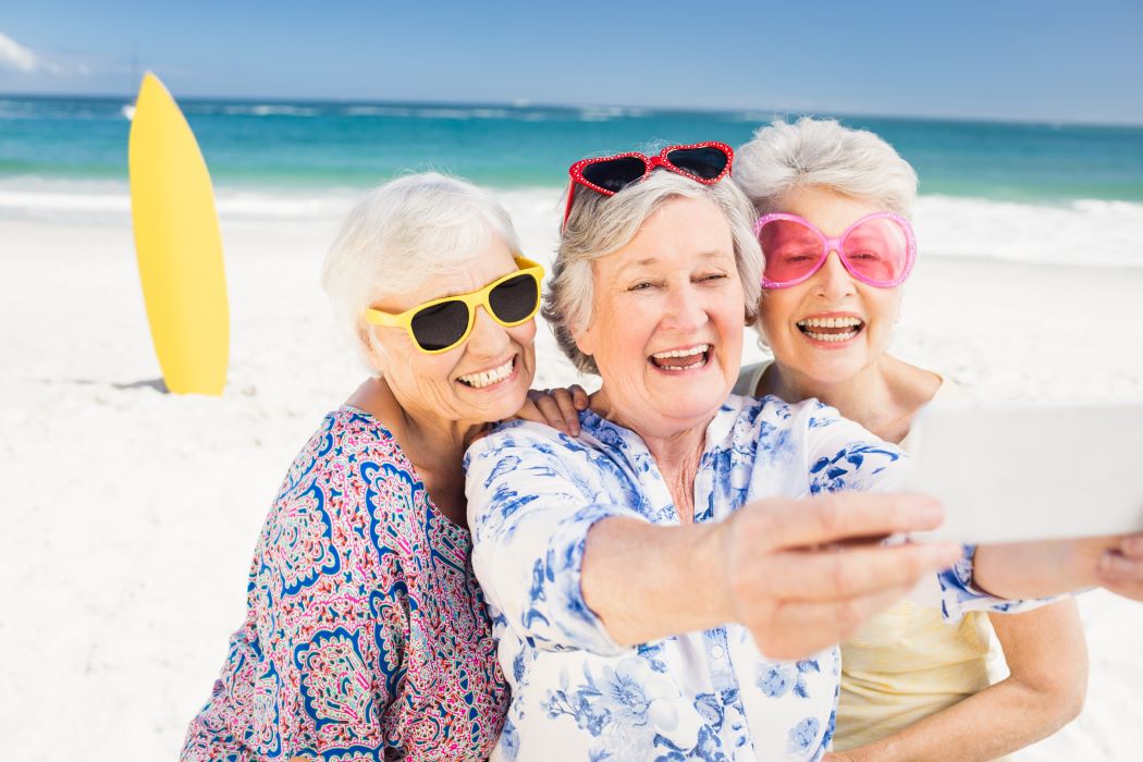 Tres persones grans amb ulleres de colors es fan un selfie a la platja, amb una taula de surf groga i el mar blau de fons