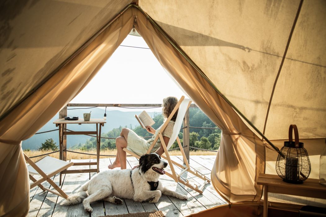 Interior de tienda de glamping con apertura triangular, terraza de madera, persona leyendo en silla y perro tumbado, paisaje de montaña al fondo y luz natural cálida