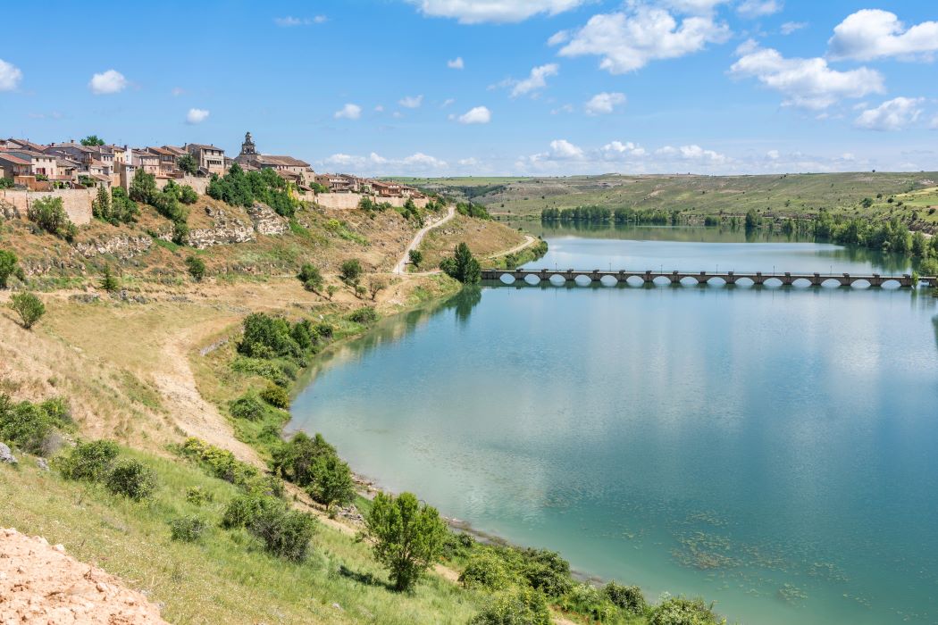 Maderuelo, pueblo en lo alto de una ladera junto a un río tranquilo con un puente de varios arcos de piedra, paisaje rural verde y cielo despejado
