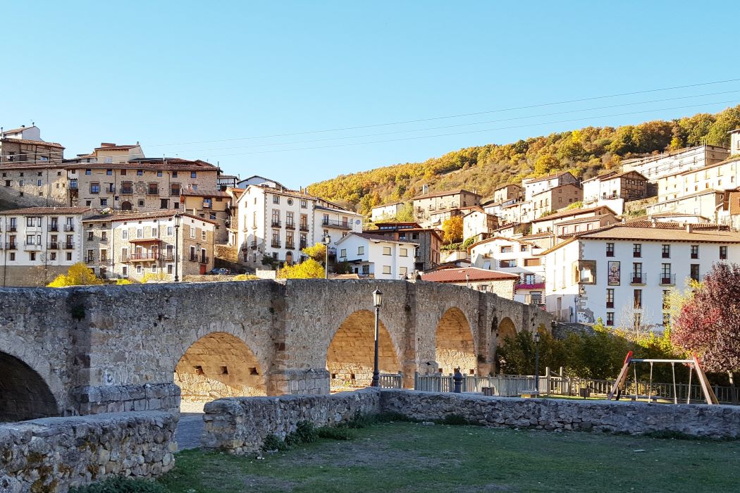 Vista del pueblo de Villoslada de Cameros, con un puente de piedra sobre un río en primer plano y casas blancas en un día soleado