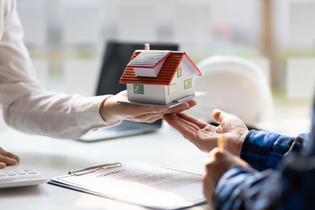 Mà sostenint una maqueta de casa lliurada a una altra persona sobre taula amb contracte i calculadora, simbolitzant l’acceptació d’una herència immobiliària