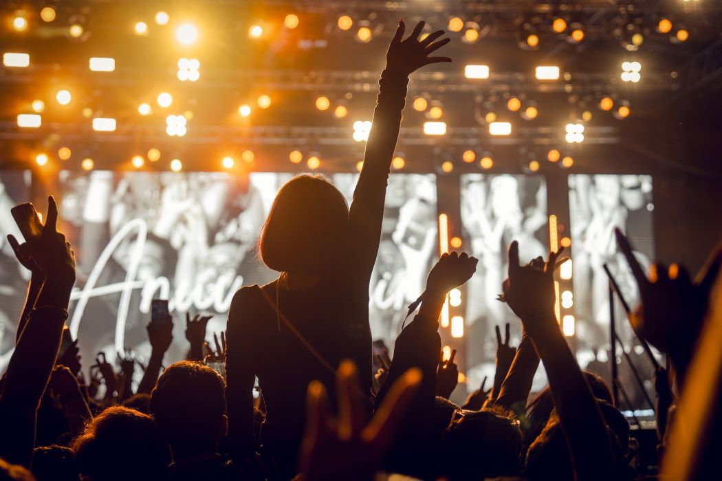 Público en concierto o festival con brazos levantados frente a un escenario iluminado con luces cálidas y pantallas, ambiente nocturno y multitud animando.